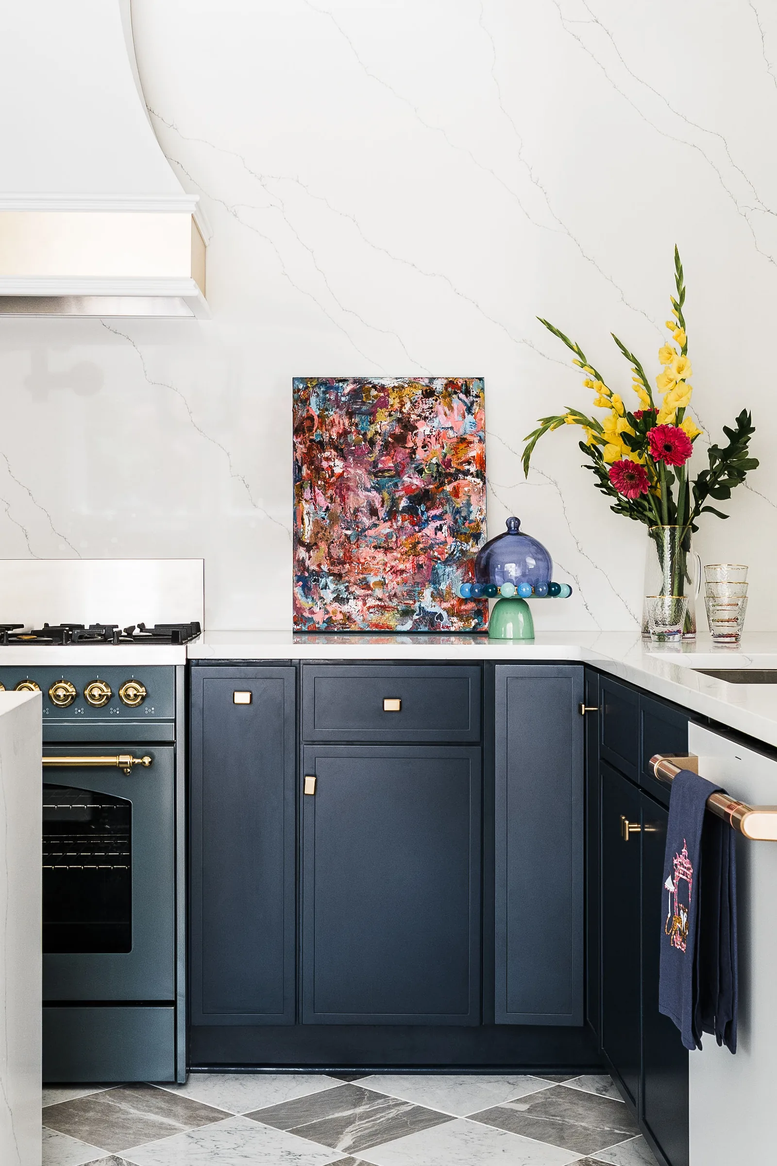 Navy cabinetry kitchen with white marble, brass hardware, abstract art, and a vase of yellow and pink flowers